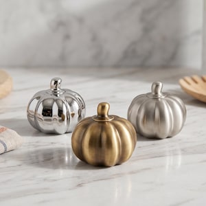 Puede incluir: Tres pequeños objetos decorativos con forma de calabaza dispuestos en grupo. Una calabaza tiene un acabado plateado pulido altamente reflectante, otra tiene un acabado de latón antiguo cepillado y otra tiene un acabado plateado cepillado mate, cada una con un pequeño tallo.