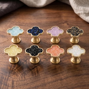 Puede incluir: Ocho pomos para armarios en forma de trébol con bases de metal en tono latón. Cada pomo presenta una incrustación central de pequeños cristales texturizados y brillantes en varios colores: azul claro, azul oscuro, morado claro, negro, amarillo, melocotón y blanco.