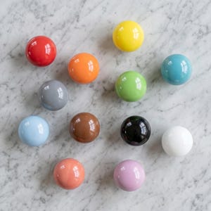 May include: A collection of twelve spherical cabinet knobs in a variety of solid colors including red, yellow, orange, light blue, gray, green, teal, brown, black, white, peach, and light purple, each featuring a smooth, glossy finish.
