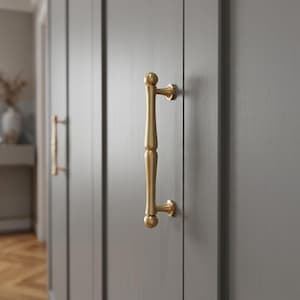 May include: A vertical brass cabinet handle with a smooth, rounded, spindle-like shape and ball-shaped ends, mounted onto a flat, grey-painted wooden door panel. The hardware features a brushed metallic finish and two circular mounting points.