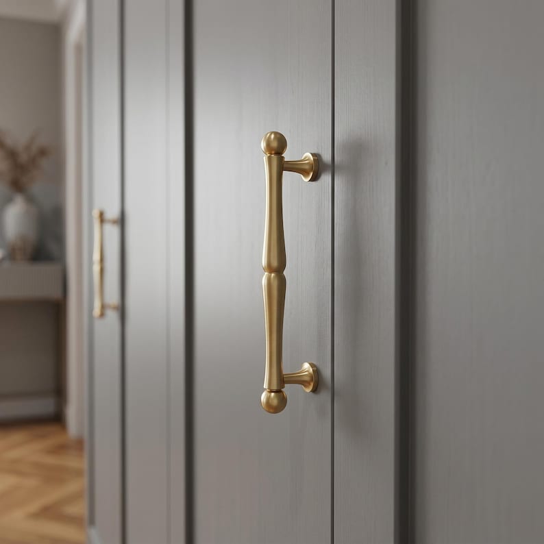 May include: A vertical brass cabinet handle with a smooth, rounded, spindle-like shape and ball-shaped ends, mounted onto a flat, grey-painted wooden door panel. The hardware features a brushed metallic finish and two circular mounting points.