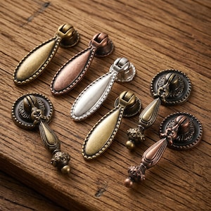 Puede incluir: Un conjunto de seis tiradores de cajón de metal con varios acabados, incluyendo latón antiguo, cobre y plata. Cada tirador presenta una pieza en forma de lágrima unida a una base circular con detalles decorativos y un mecanismo de bisagra.