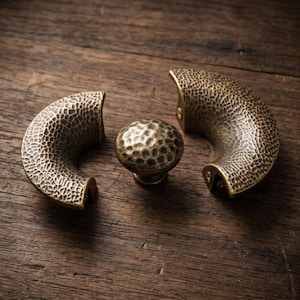 May include: Two crescent-shaped cabinet pulls and one round cabinet knob, all featuring a dark bronze finish with a textured, hammered surface. The hardware pieces are arranged together, showcasing the consistent dimpled metal pattern throughout.