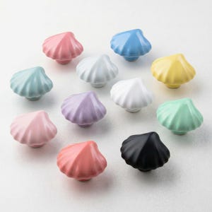 May include: A collection of eleven ceramic cabinet knobs shaped like seashells, each featuring a distinct solid color including pink, light blue, white, yellow, lavender, mint green, coral, and black, arranged in a scattered pattern.