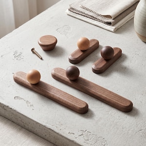 May include: A collection of wooden cabinet hardware including two long rectangular pulls with spherical knobs, two shorter rectangular pulls with spherical knobs, one circular knob, and a single metal screw. The wood features light and dark tones.