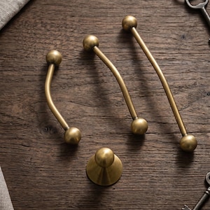 May include: Three curved brass cabinet pulls with spherical ends and one matching circular brass knob. The hardware features a smooth, matte metallic finish. The items are arranged on a dark wood surface alongside two metal keys.