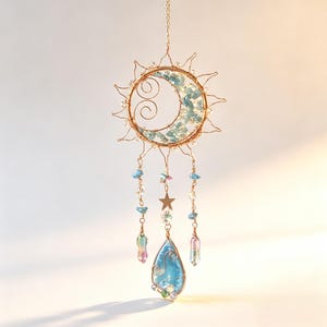 May include: A hanging sun and moon ornament crafted from gold-toned wire. The circular frame features a crescent moon filled with light blue stone chips and wire swirls. Three dangling strands include clear beads, blue stones, a metal star, and a teardrop pendant.
