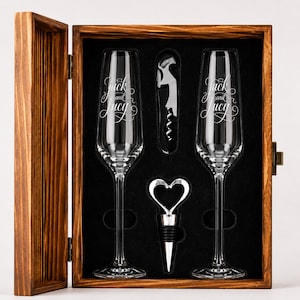 May include: A wooden gift box containing two clear glass champagne flutes, a metal corkscrew, and a heart-shaped bottle stopper. The flutes are etched with the text Jack and Lucy in a cursive script. The interior is lined with black foam.