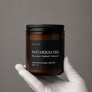 Könnte beinhalten: Ein braunes Glas-Kerzenglas mit schwarzem Deckel und schwarzem Etikett. Auf dem Etikett steht "PATCHOULI VEIL" mit Duftnoten. Die Kerze ist handgegossen und aus Sojawachs hergestellt, mit einem Gewicht von 200 g. Das Glas wird von einer behandschuhten Hand gehalten.