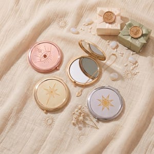 Può includere: Quattro specchietti da borsa rotondi con finiture in oro rosa, oro e argento, caratterizzati da disegni celesti incisi tra cui un occhio e stelle. Uno specchio dorato è aperto, rivelando una superficie riflettente, con una catenina dorata.
