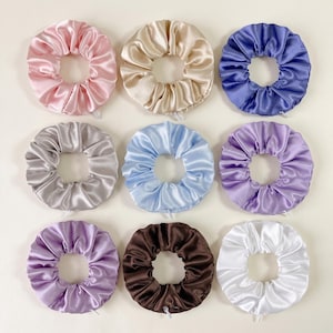 Puede incluir: Una colección de nueve coleteros circulares dispuestos en una cuadrícula de tres por tres. Los coleteros presentan una tela suave y brillante en varios colores, incluyendo rosa claro, beige, morado, gris, azul claro, lavanda, marrón oscuro y blanco.