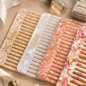 May include: Four rectangular hair combs made of marbled acrylic in shades of beige, white, and pink. Each comb features a unique name engraved in gold cursive script: Sophia, Isabelle, Ava, and Sophia. The combs have wide, evenly spaced teeth.