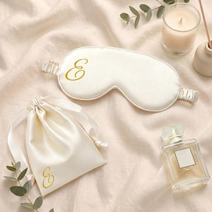 Könnte beinhalten: Eine cremefarbene Schlafmaske aus Satin und ein passender Beutel mit Kordelzug, beide mit einem goldenen, kursiven Buchstaben E versehen. Die Schlafmaske hat ein elastisches Band und der Beutel besteht aus glattem Stoff mit passenden Bändern.