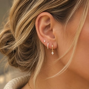 May include: A set of three gold-toned earrings worn in a single ear. The set includes a small star-shaped stud, a thin hoop earring encrusted with tiny clear stones, and a gold hoop earring featuring a dangling, diamond-shaped clear stone charm.