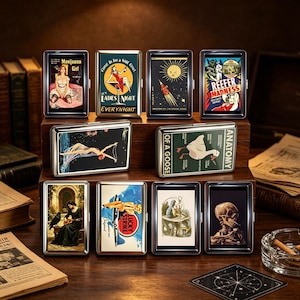May include: A collection of ten rectangular metal cigarette cases featuring various vintage-style graphic prints, including a skeleton, a goose anatomy diagram, a rocket, and pin-up illustrations, arranged on a tiered wooden display stand.