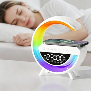 May include: A white crescent-shaped alarm clock with a multi-colored LED light strip along the inner curve. The device features a digital display showing 23:00, control buttons on the top, and a flat surface for wireless smartphone charging.