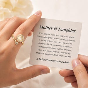 May include: A gold-colored ring with a round, light-colored stone surrounded by small clear stones. The ring is on a finger, next to a card with the text "Mother & Daughter" and a poem about the bond between a mother and daughter.