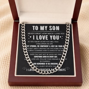 Puede incluir: Un collar de cadena plateada exhibido en una caja de madera oscura. El collar se encuentra sobre una tarjeta negra con texto blanco que dice "TO MY SON" y un mensaje cariñoso. La caja está abierta, mostrando el collar y la tarjeta.