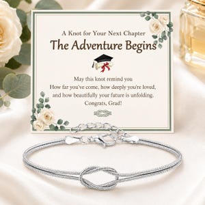 May include: A silver-toned snake chain bracelet featuring a central knot design and a lobster claw clasp. The bracelet is displayed in front of a rectangular card with floral accents, a graduation cap icon, and the text A Knot for Your Next Chapter The Adventure Begins May this knot remind you How far you've come, how deeply you're loved, and how beautifully your future is unfolding. Congrats, Grad!