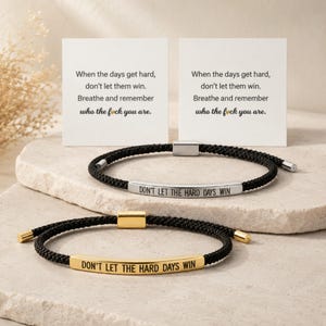 Puede incluir: Dos pulseras de cuerda negra con barras metálicas grabadas con "DON'T LET THE HARD DAYS WIN". Las pulseras se muestran con dos tarjetas que dicen "Cuando los días se pongan difíciles, no dejes que ganen. Respira y recuerda quién eres."