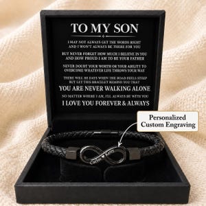 May include: A black braided leather bracelet with a metal infinity symbol centrepiece featuring the engraved text 'Carlos Ketty' with a heart icon, presented in a black gift box with a printed message card addressed 'TO MY SON'.