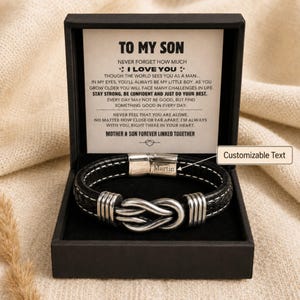 Puede incluir: Pulsera de cuero negro con un diseño de nudo infinito plateado, presentada en una caja de regalo negra. La caja contiene un mensaje impreso: "TO MY SON, I LOVE YOU..." El cierre de la pulsera está grabado con el nombre "Martin".