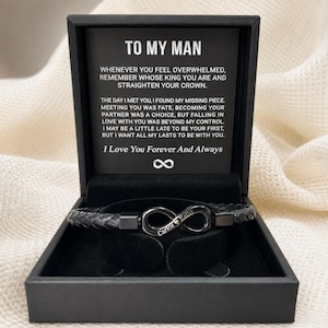 Op de afbeelding: Zwart gevlochten leren armband met een oneindigheidssymbool sluiting, gepresenteerd in een zwarte geschenkdoos. Het oneindigheidssymbool is gegraveerd met de namen "Carlos & Kelly". De doos bevat de tekst "To My Man".