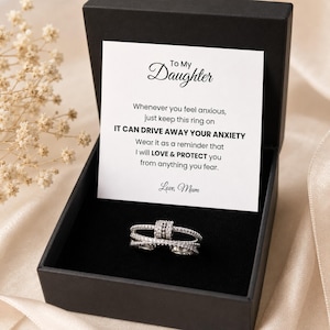 Könnte beinhalten: Ein silberner Ring mit mehreren Bändern und kleinen, klaren Steinen, präsentiert in einer schwarzen Geschenkbox. Die Box enthält eine Karte mit dem Text "To My Daughter" und eine Botschaft über Liebe und Schutz.
