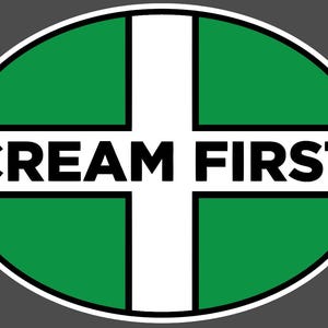 May include: An oval sticker featuring a green background divided by a white cross. The text CREAM FIRST is printed in bold black capital letters across the horizontal bar of the cross. The entire design is enclosed by a thin white border.