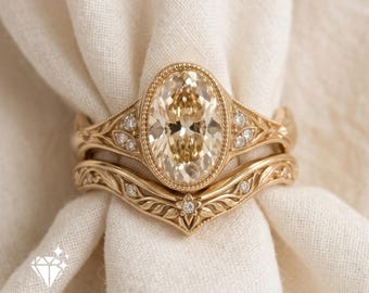 May include: A gold ring set featuring an oval-cut champagne-colored center stone surrounded by a beaded border and small accent diamonds. The band and matching curved wedding band display intricate leaf engravings and metalwork.
