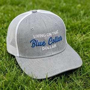 Może przedstawiać: Szara czapka typu trucker z białą siatką z tyłu i zakrzywionym daszkiem. Czapka ma wyhaftowany tekst "LIVING ON THAT Blue Collar DOLLAR" w kolorze niebieskim. Czapka leży na zielonej trawie.