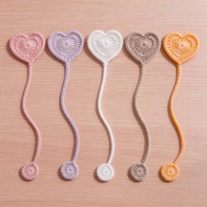 Puede incluir: Cinco marcapáginas de ganchillo, cada uno con una parte superior en forma de corazón y una pequeña base circular conectadas por un cordón largo y fino. El conjunto incluye un marcapáginas de cada color: rosa claro, lavanda, blanco, beige y naranja.