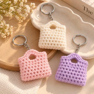 May include: Three miniature crochet bag keychains in cream, light pink, and lavender. Each keychain features a textured, hand-stitched pattern with a small integrated handle and a silver-toned metal split ring attached by a short chain link.