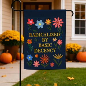 Peut inclure: Un drapeau de jardin rectangulaire bleu marine foncé avec le texte jaune "RADICALIZED BY BASIC DECENCY" centré sur le tissu. Le motif est entouré de diverses illustrations florales colorées dans des tons de rose, orange et bleu clair.