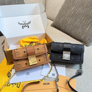 Könnte beinhalten: Zwei rechteckige Umhängetaschen mit MCM-Logomuster. Eine Tasche ist in Cognacbraun mit goldfarbenen Beschlägen und goldener Kette. Die andere ist schwarz mit silberfarbenen Beschlägen und silberner Kette. Beide haben einen Überschlag.