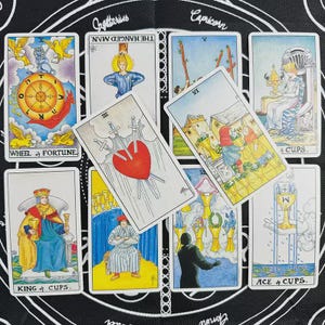 Könnte beinhalten: Eine Sammlung von Tarotkarten, die auf einer schwarzen Oberfläche mit weißen Tierkreiszeichen angeordnet sind. Zu den sichtbaren Karten gehören das Rad des Schicksals, der Gehängte, die Drei der Schwerter, die Sechs der Kelche und der König der Kelche.