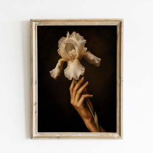 Op de afbeelding: Een ingelijste kunstdruk met een crèmekleurige irisfleur die door een hand wordt vastgehouden. De donkere achtergrond contrasteert met de bloem en de hand. De lijst is licht en neutraal.
