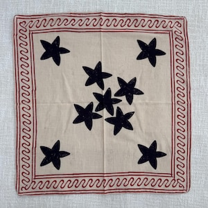 May include: A square fabric handkerchief in a light beige color featuring a central cluster of six black stars and four additional black stars in the corners. The border is decorated with a continuous red wavy line pattern on a square frame.
