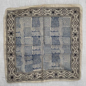 May include: A square beige bandana featuring a dark blue and black block-printed pattern. The design consists of a central grid of alternating geometric shapes, including squares, triangles, and wavy lines, framed by a floral and diamond border.