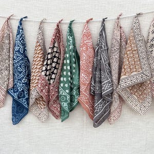 May include: Ten square fabric bandanas hanging on a white string. Each piece features a unique block-printed pattern in various colors including blue, green, terracotta, grey, and black on a light cream background with decorative border designs.