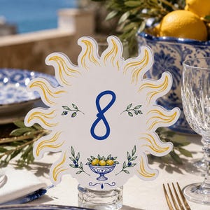 Puede incluir: Una tarjeta de número de mesa troquelada con forma de sol con rayos amarillos, que presenta un gran número 8 azul en el centro. El diseño incluye ramas de olivo verdes y un pequeño cuenco azul lleno de limones amarillos en la parte inferior.