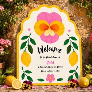 May include: A scalloped-edge sign with a yellow border featuring a large pink and yellow pansy at the top, two yellow lemons on green leafy branches, and a small pink flower at the bottom. The text reads Welcome to the Bridal shower of Violet, 12 July 2025, Impruneta, Florence, Formal invitation to follow.