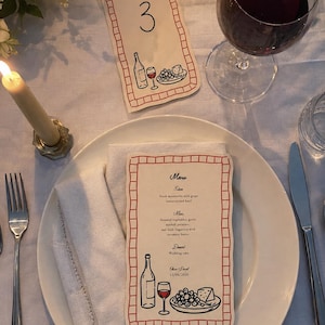 May include: A white dinner plate topped with a folded white napkin and a rectangular menu card featuring a red grid border, a wine bottle, a glass of red wine, and a plate of grapes and cheese. The menu text lists courses and the date 23/06/2030.