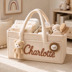 Peut inclure: Un panier de rangement en tissu texturé couleur crème avec deux anses, portant le prénom Charlotte brodé en lettres cursives marron. Le panier comprend une breloque ours en peluche, un écusson tête de lion et des compartiments internes.