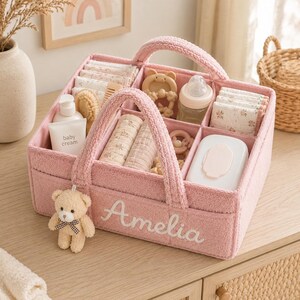 Puede incluir: Una cesta organizadora para bebé de tela texturizada rosa con dos asas, que presenta el nombre Amelia bordado en letra blanca en la parte frontal. Un pequeño oso de peluche cuelga de un lado. El interior está dividido en compartimentos con crema para bebé, un biberón, paños doblados y un recipiente blanco para toallitas.