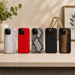 May include: Five rectangular smartphone cases arranged in a row, each featuring a distinct textured finish. The designs include white, solid red, grey and black snakeskin pattern, solid black, and a brown and black snakeskin pattern, all with camera cutouts.