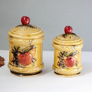 May include: Two ceramic canisters with lids. The canisters are tan with fruit designs, including apples and grapes. Each lid is topped with a red apple. The canisters are of different sizes and are likely for kitchen storage.