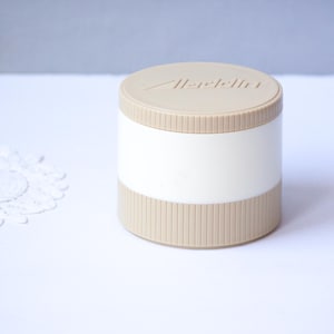 May include: A cylindrical container with a beige lid and base, and a white central band. The lid is embossed with the word "Aladdin". The base has vertical ribbed detailing. The container sits on a white surface.