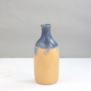 May include: A handcrafted ceramic vase with a rounded body and a narrow neck. The vase features a two-tone design, with a light orange base and a blue top. Blue drips cascade down the sides, adding a unique touch. This decorative vase is suitable for home decor.