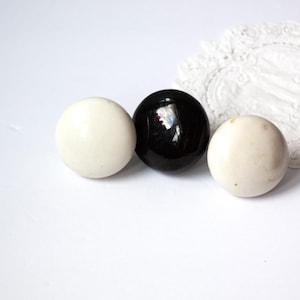 May include: Three round knobs in a row, two are off-white and one is glossy black. A white, lacy doily is in the background. The knobs are likely for furniture or decorative purposes.